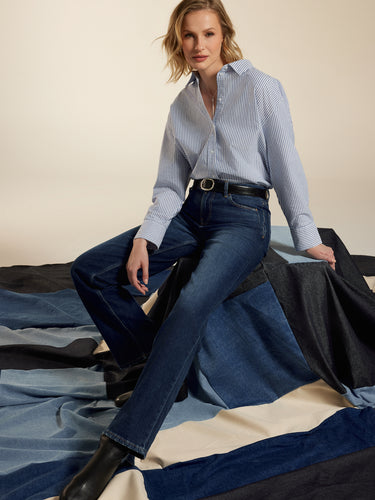 Woman wearing a light blue striped shirt and dark jeans from Cup of Joe Denim sitting on a bed with striped bedding.