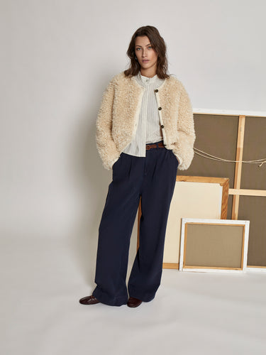 Woman wearing a beige teddy jacket and navy pants from Peppercorn standing in front of framed artwork.
