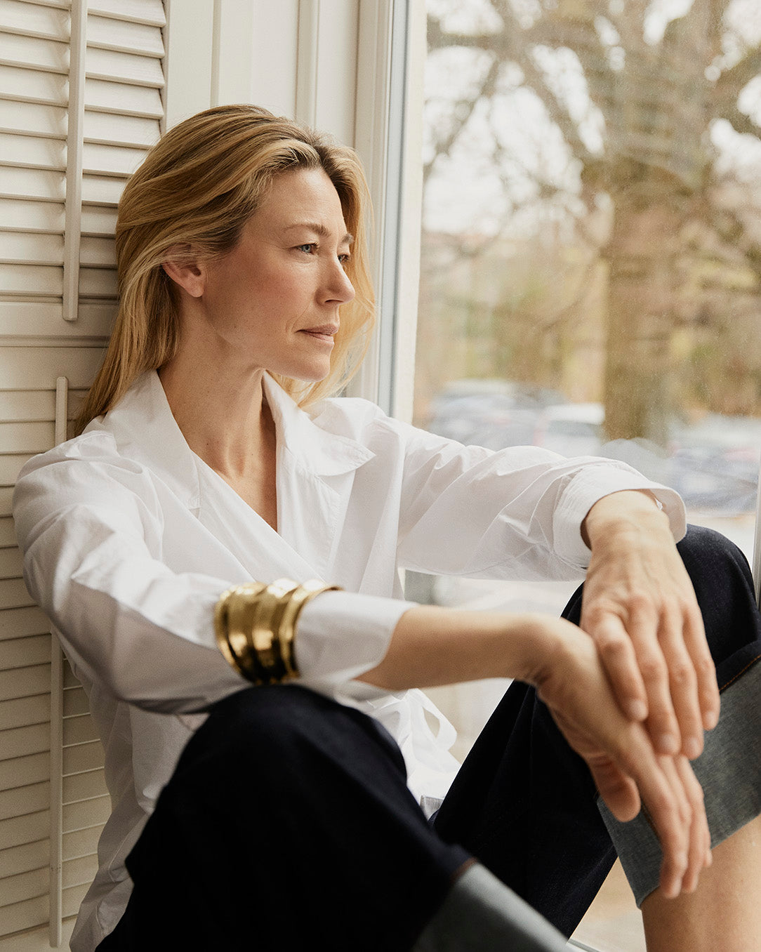 Woman in a white shirt  and dark denim jeans from Masai Copenhagen sitting by a window with a blurred outdoor background