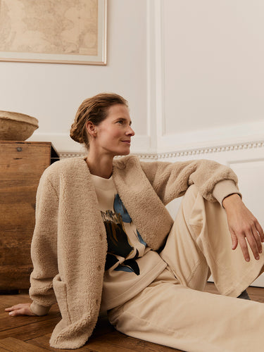 Woman sitting on the floor wearing a beige Masai cardigan and pants in a room with wooden furniture and a map.