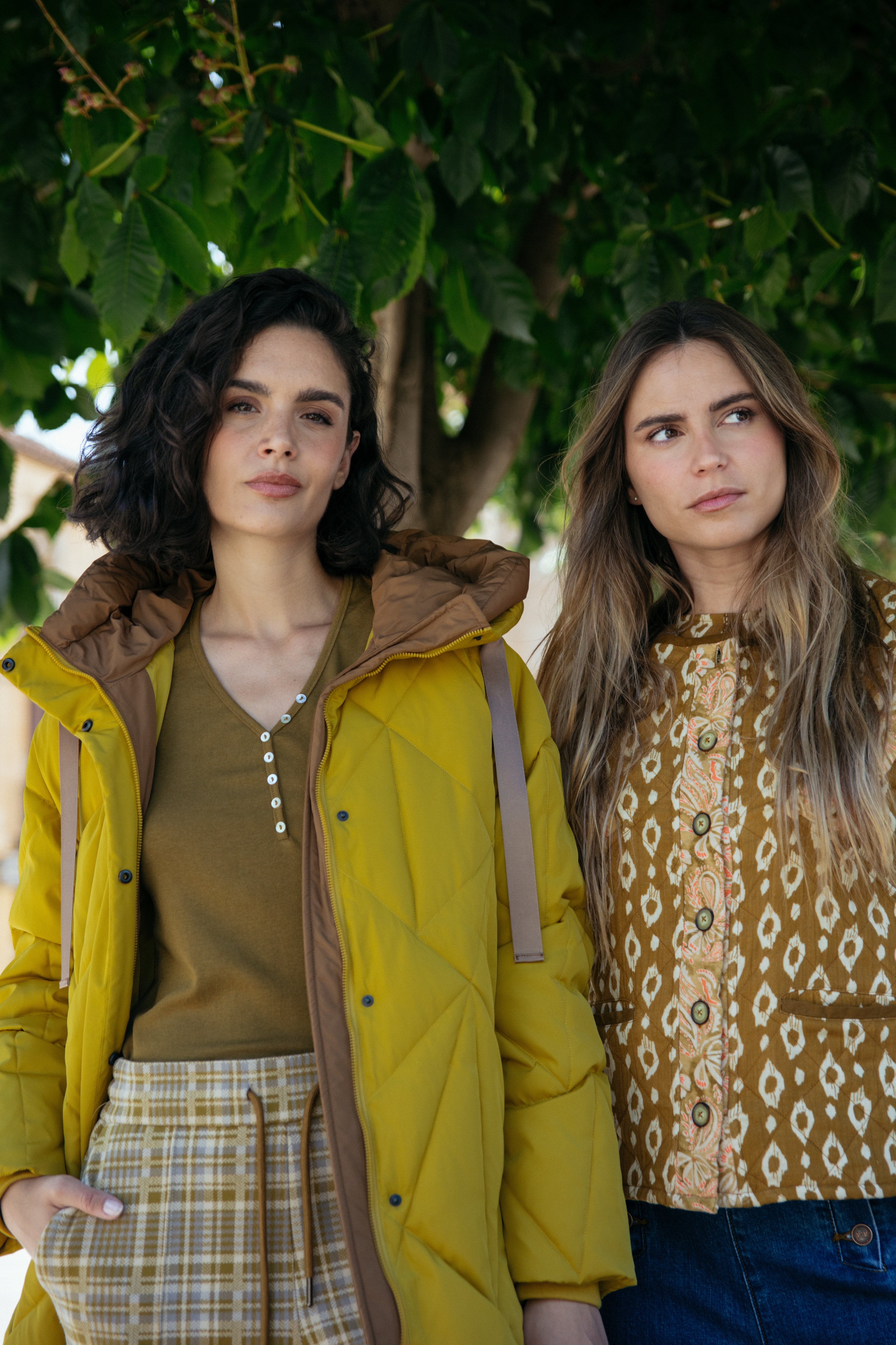 Two women standing outdoors under a tree, one wearing a yellow raincoat and plaid skirt, the other in a patterned top from MD'M fall winter 2025